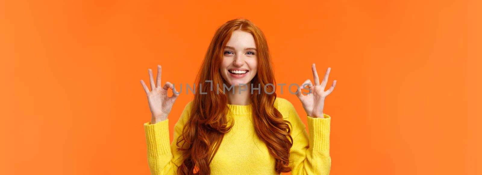 Fashion, shopping, winter holidays concept. Attractive cheerful redhead woman showing okay gestures and smiling, everything alright, approve or like product, recommend online store, orange background.