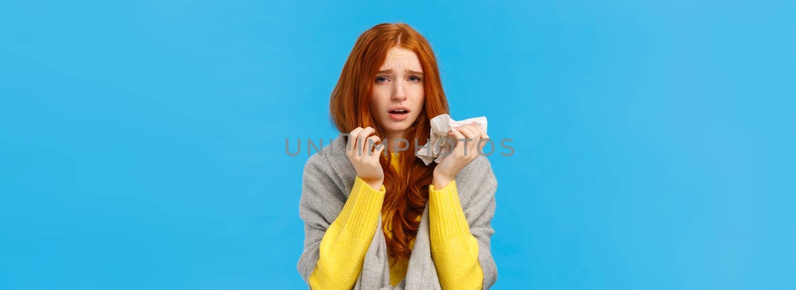 Dizzy, uneasy sad redhead female wearing scarf to get warm, catching cold, sneezing and having runny nose, using napkin, looking camera with tired exhausted expression, feel fatigue, blue background.