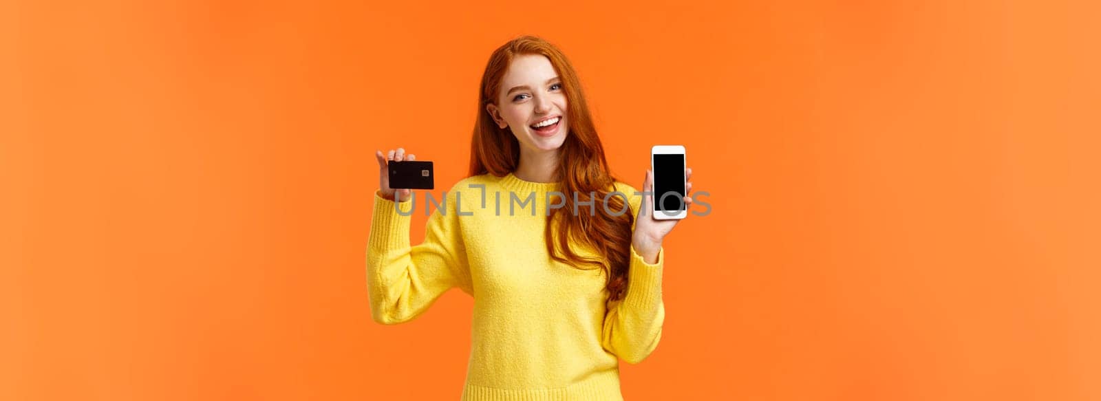 Shopping, online and finance concept. Cheerful young redhead female opened bank account, holding smartphone and credit card, smiling, recommend use online shopping application.