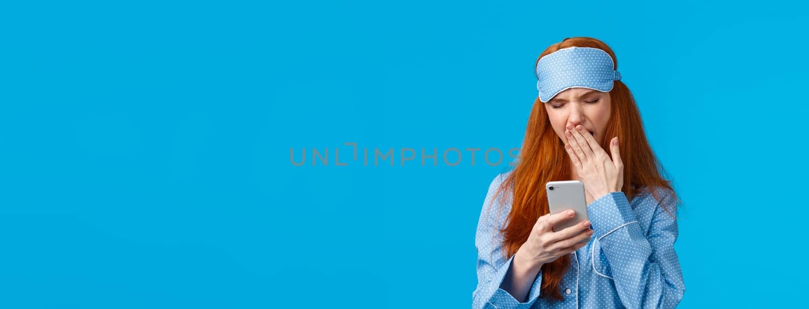 Gadgets addication, lifestyle and beauty concept. Lovely redhead girl going sleep and scrolling feed smartphone, yawning squinting feeling tired, wearing pyjama and sleep mask, blue background by Benzoix