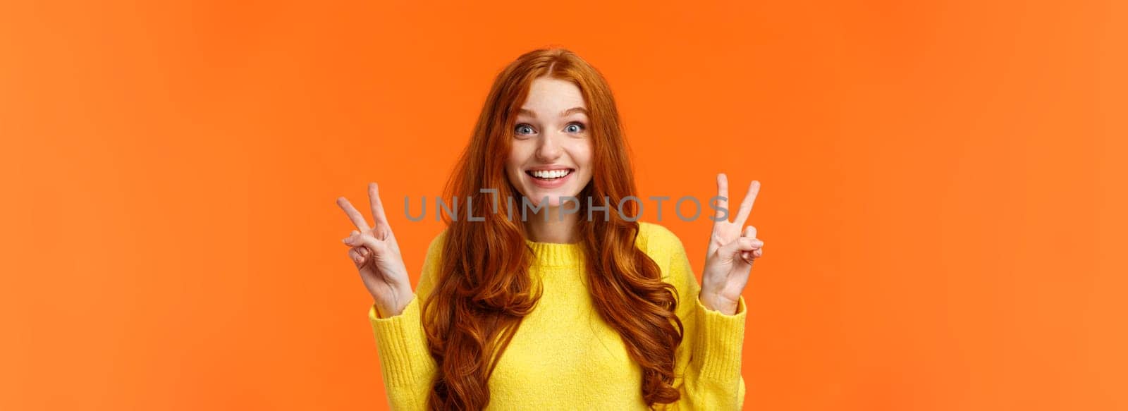 Waist-up portrait lovely and cheerful pretty redhead woman showing peace gestures, smiling camera, travel abroad during winter holidays posing near sightseeing, orange background.