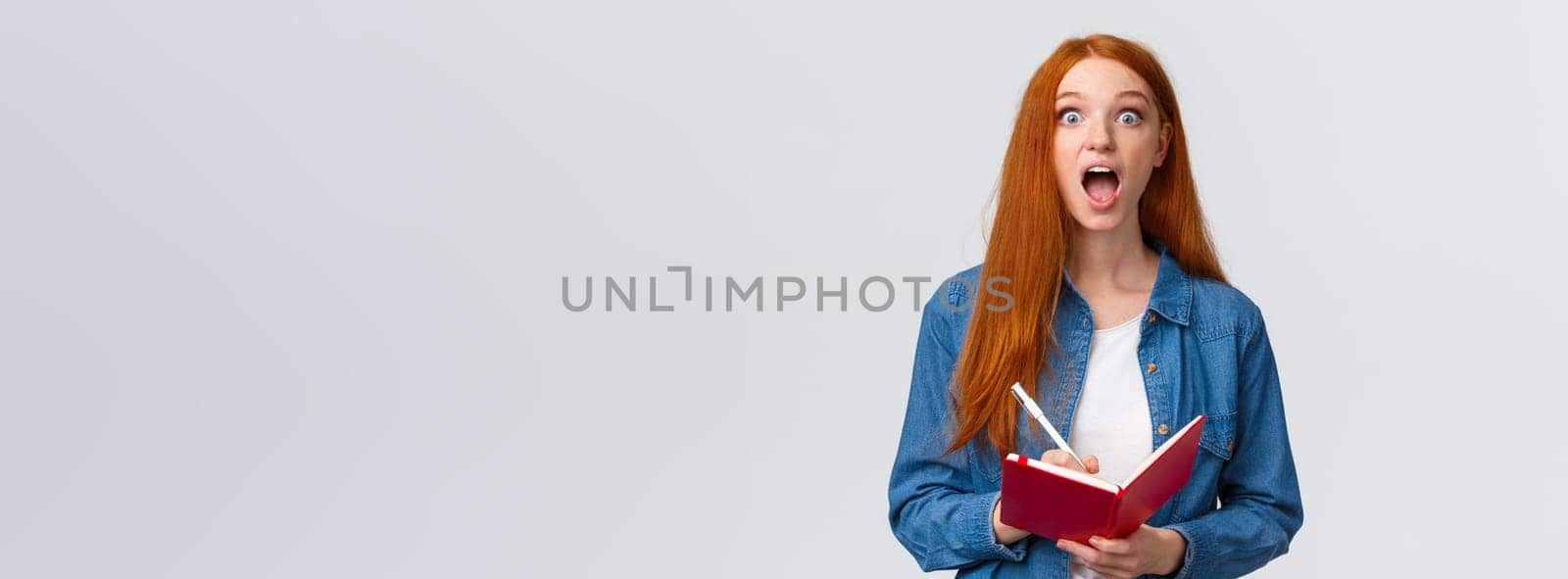 Amused and excited, astonished redhead girl fascinated with amazing lecture giving speech, writing down useful notes, holding notebook and staring thrilled camera, white background.