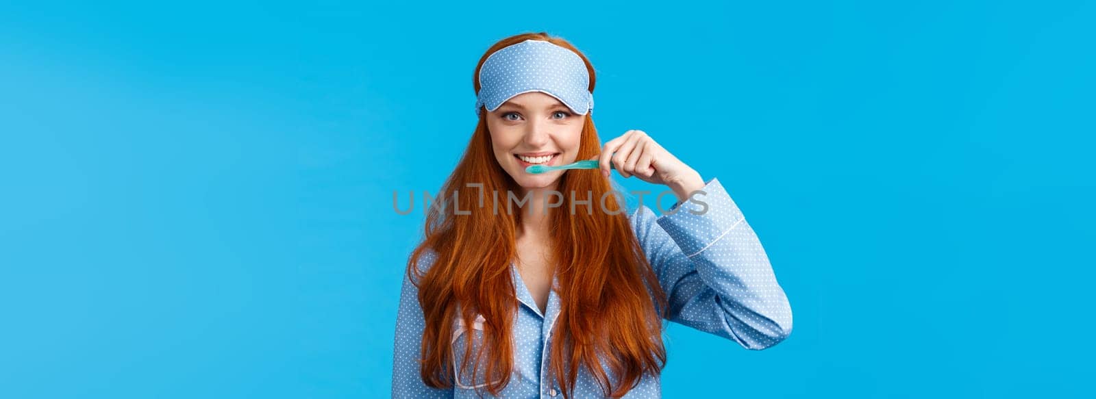 Habits, hygiene and lifestyle concept. Cute feminine redhead european woman in nightwear, sleep mask smiling and brushing teeth with toothbrush, standing blue background.