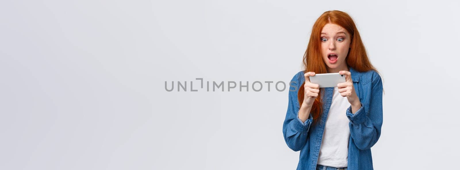 Astonished startled cute redhead woman just won challenge in awesome game, holding smartphone horizonally, playing racing, stare display amazed, cant belive she lucky, white background.