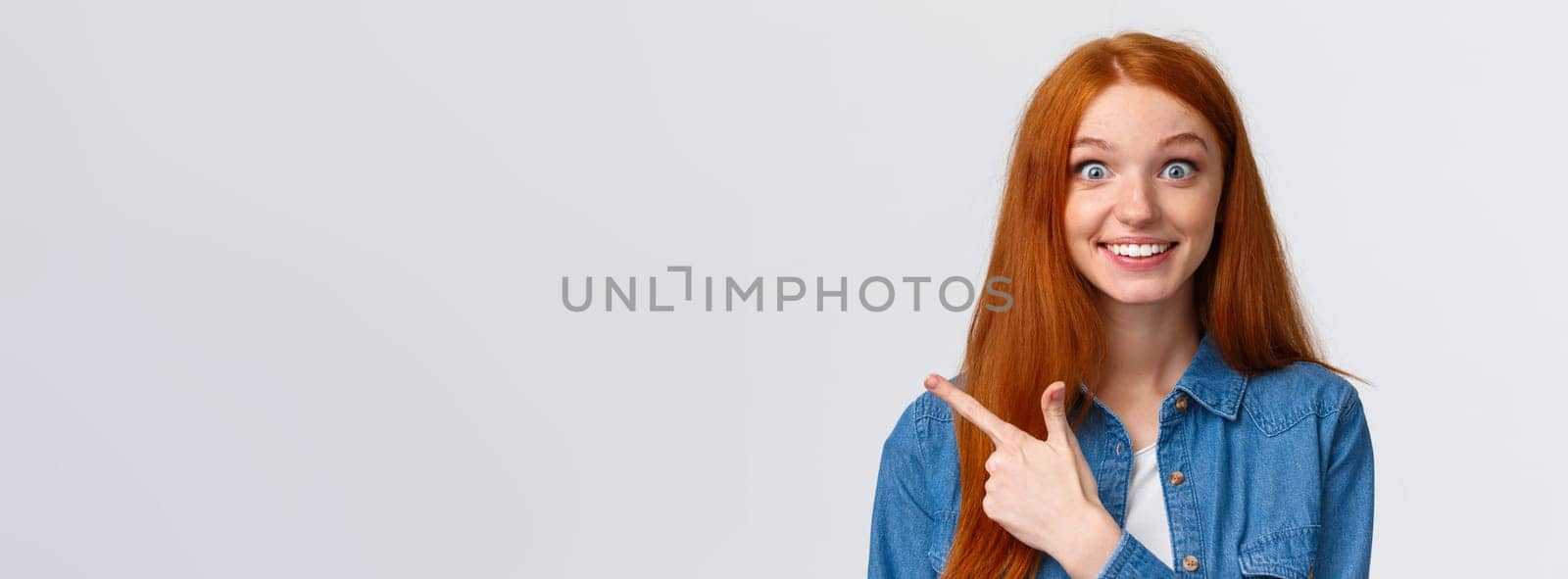 Excited and impressed, curious thrilled smiling redhead girl with long foxy, ginger hair and blue eyes, stare amused camera with wondered grin, pointing finger left, found out awesome news.