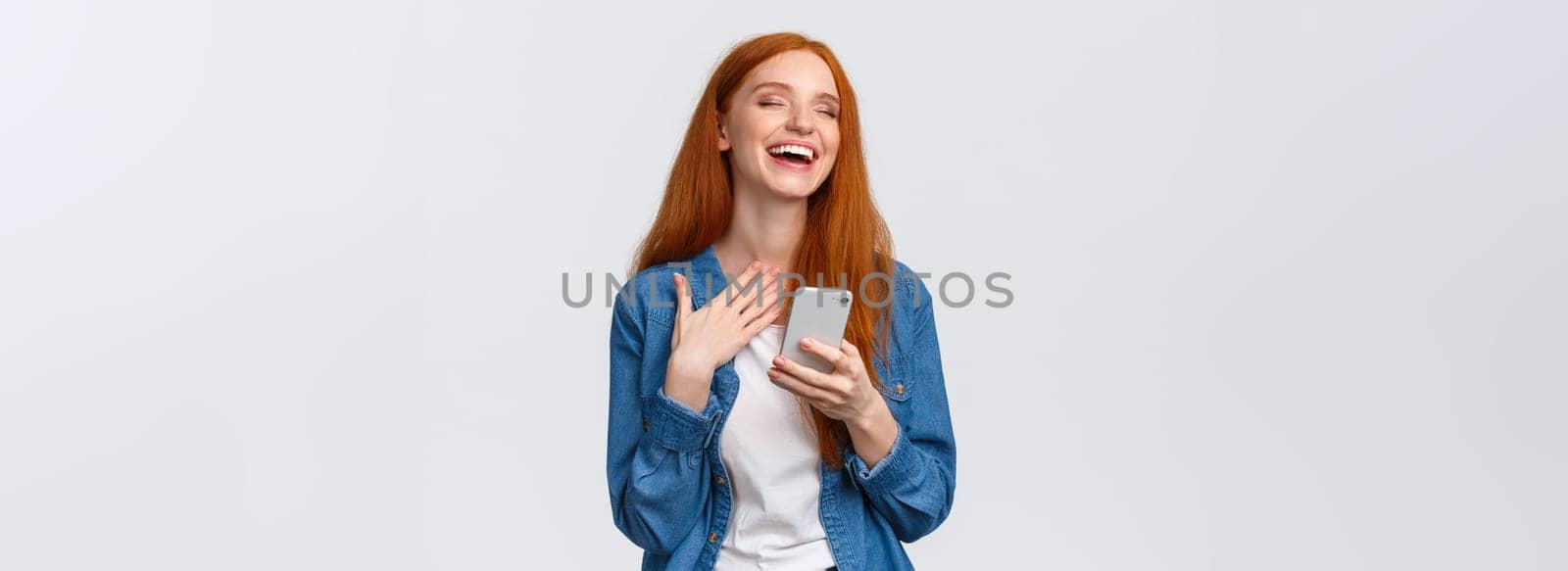 Waist-up portrait carefree and joyful, alluring redhead caucasian girl receive funny meme message, holding smartphone, laughing and close eyes dreamy, standing white background.