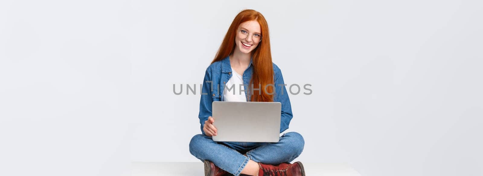 Cheerful sassy redhead female student, college girl in glasses, sitting on crossed legs with laptop, working freelance, prepare project awat from office, remote over white background.