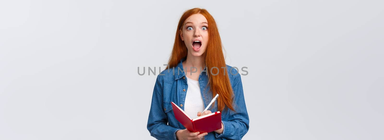 Amused and excited, astonished redhead girl fascinated with amazing lecture giving speech, writing down useful notes, holding notebook and staring thrilled camera, white background.