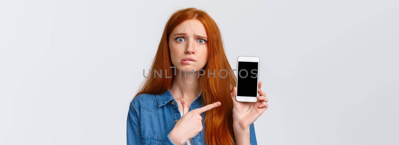 Worried, insecure and alarmed cute redhead girl asking for advice having problems messaging with stranger on dating app, dont know what answer standing nervous pointing finger at smartphone display.