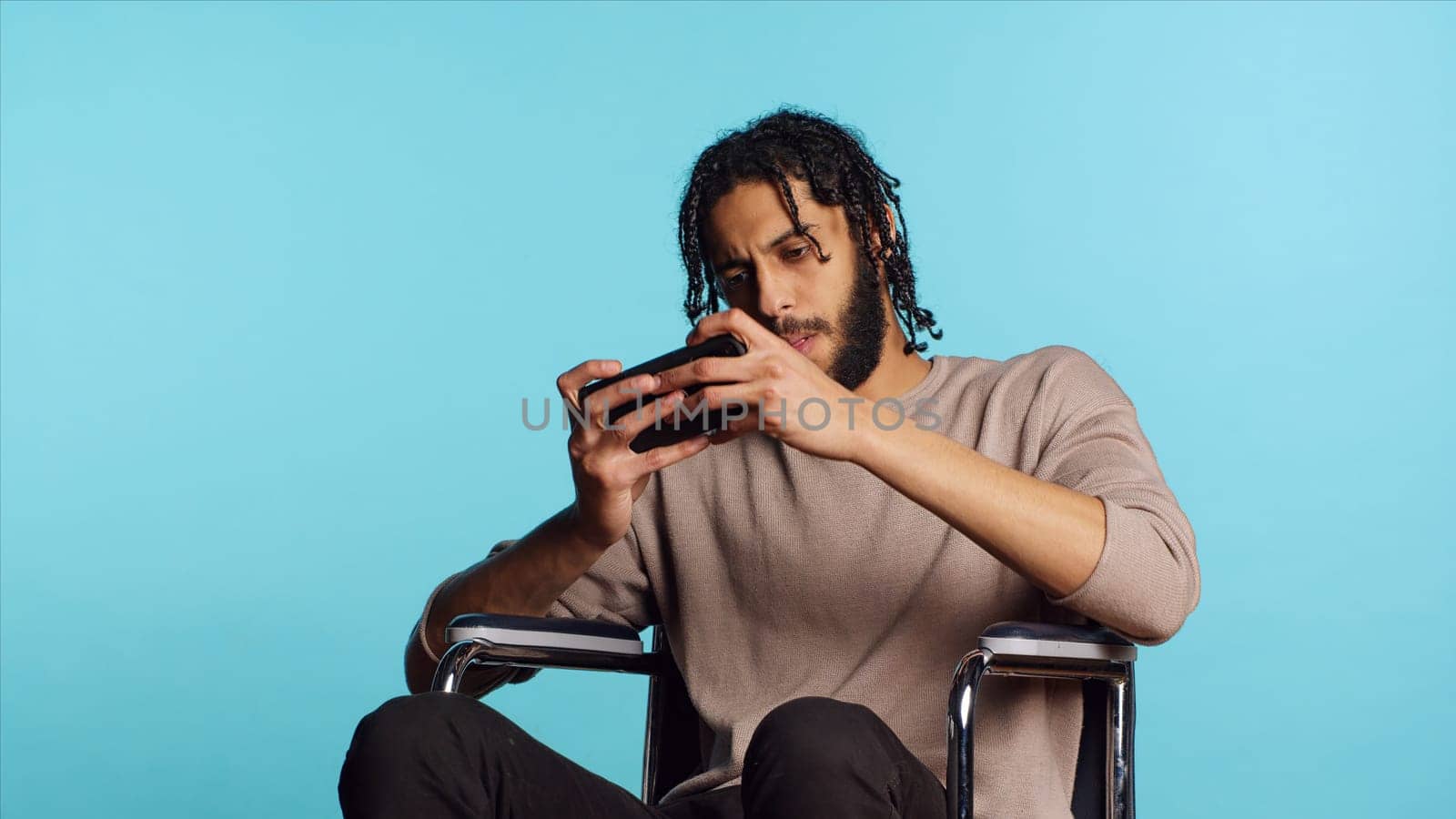 Man with disability in wheelchair playing videogames on cellphone, enjoying leisure time. Middle Eastern gamer enjoying game on mobile phone, having fun, studio background, camera A