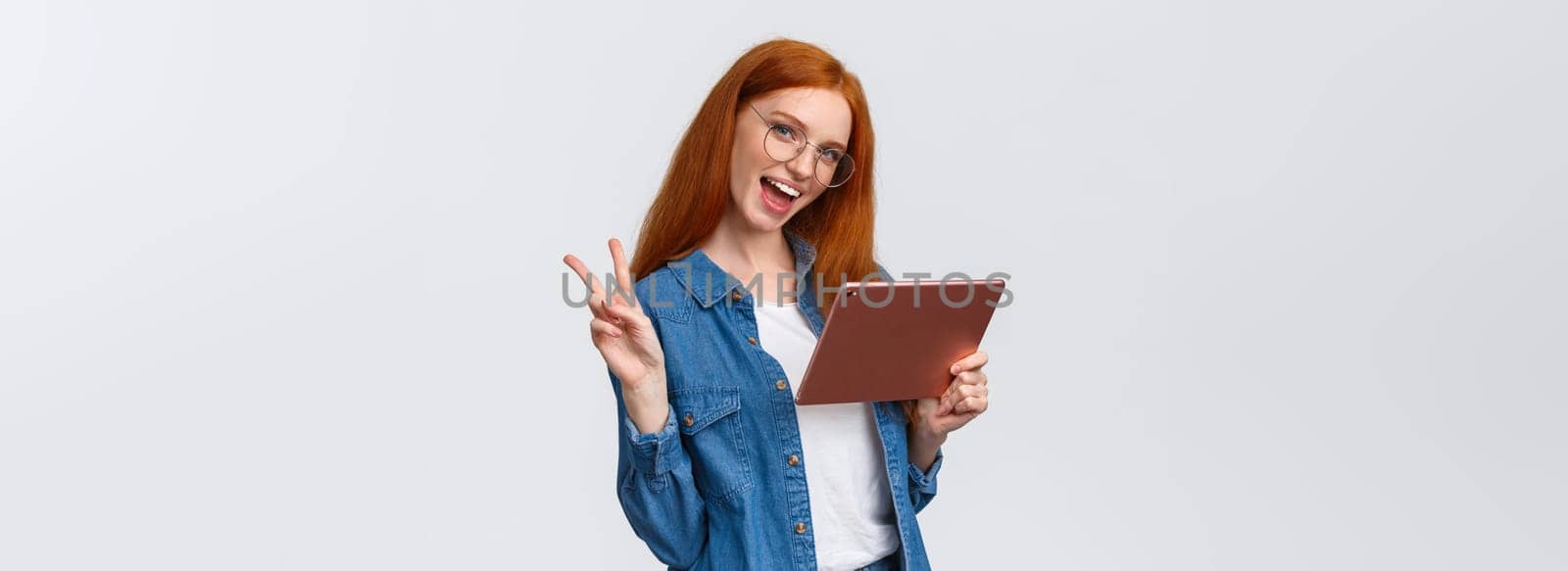 Sassy and cute, carefree happy redhead woman in glasses, denim shirt, showing peace sign and smiling camera cheerful as holding digital tablet, watching vlog online, working from distance.