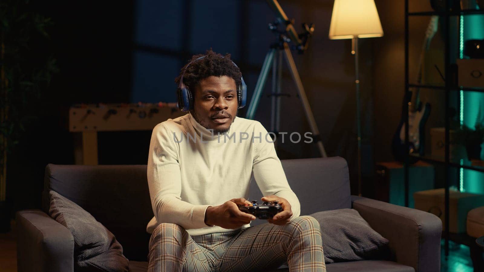 Man in rgb lit apartment playing video games on couch, enjoying day off from work. Gamer battling enemies in online multiplayer shooter on gaming console, talking with teammates through headphones