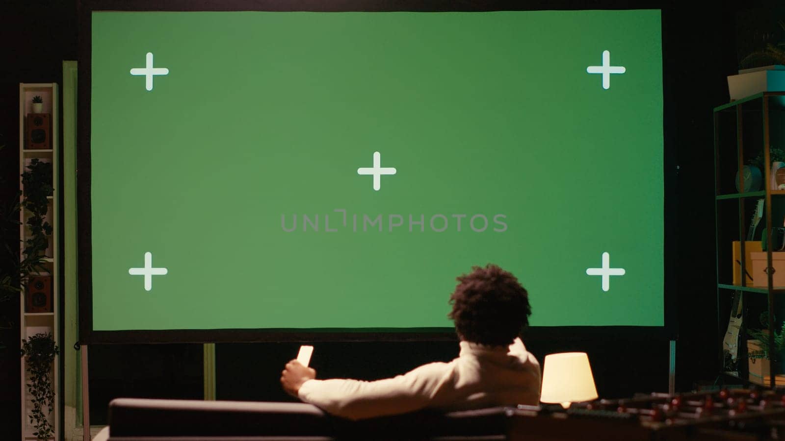 African american man using video projector to binge TV series on subscription based streaming services. Cord cutter enjoying VOD shows on gigantic green screen television set