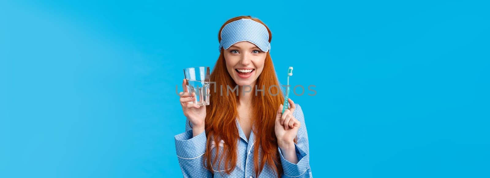 Advertising, hygiene and people concept. Feminine cute redhead girl feeling upbeat and enthusiastic start day right, drinking water, holding glass and toothbrush, wear nightwear and sleep mask.