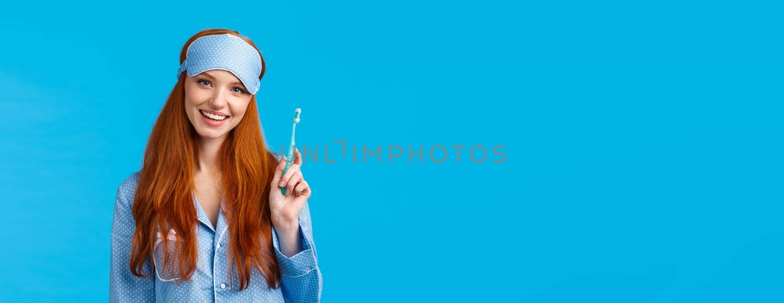 Health, beauty and lifestyle concept. Cheerful pretty european woman with blue eyes, toothy smile, holding toothbrush and smiling delighted, wearing sleep mask, nightwear, blue background.