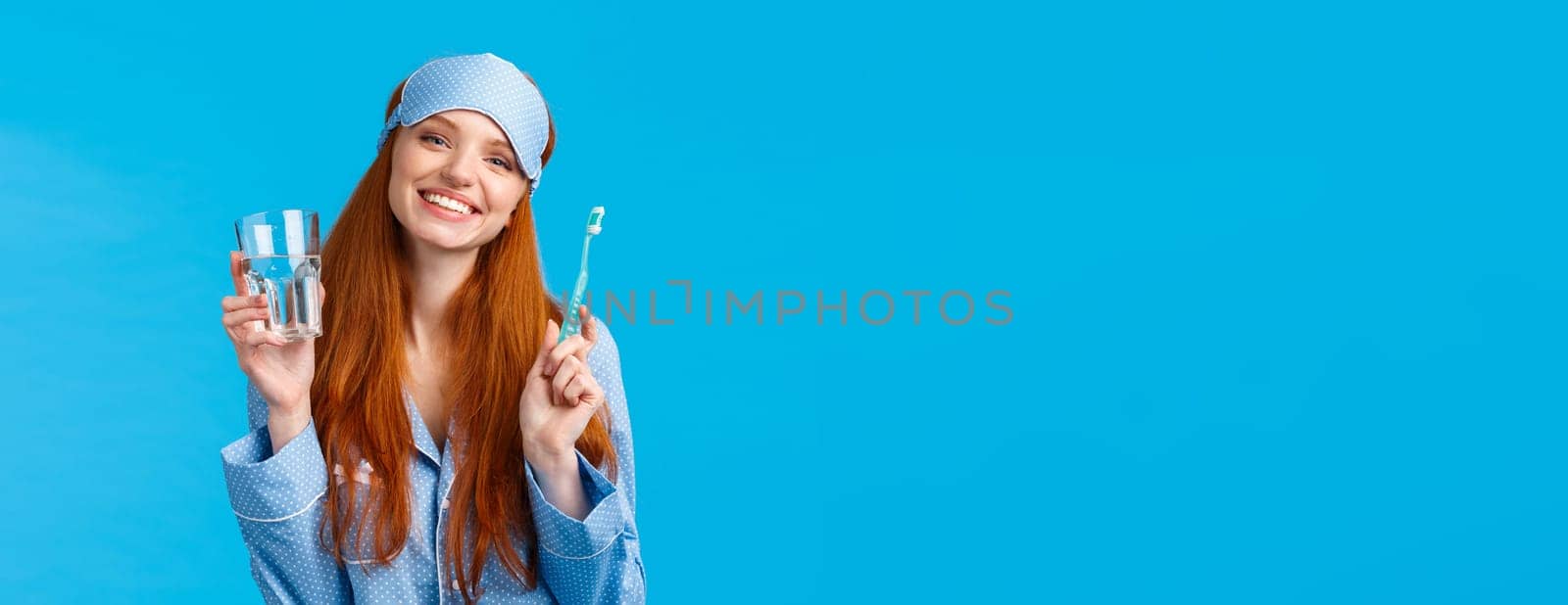 Happy smiling and carefree redhead enthusiastic girl in sleep mask, pyjama, holding glass water and toothbrush, taking care mouth hygiene, dental daily routine, blue background.