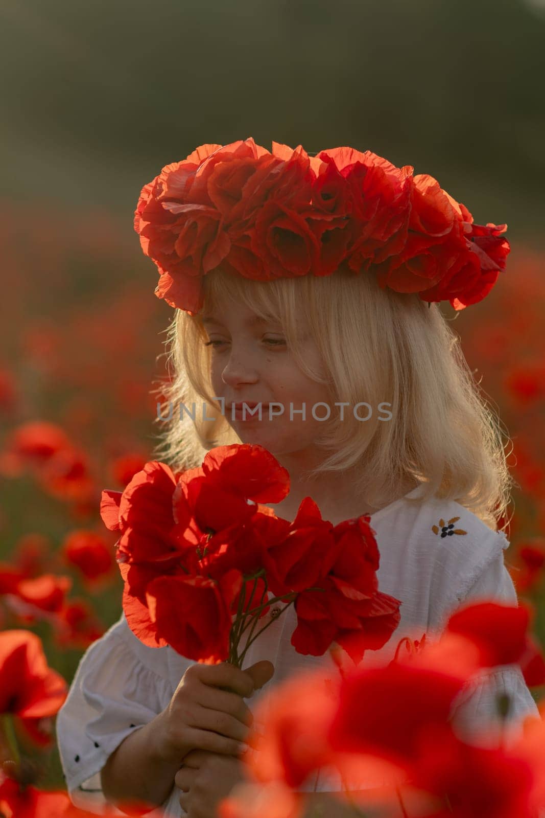 A young girl wearing a red flower crown is standing in a field of red poppies. She is holding a bouquet of red flowers in her hand