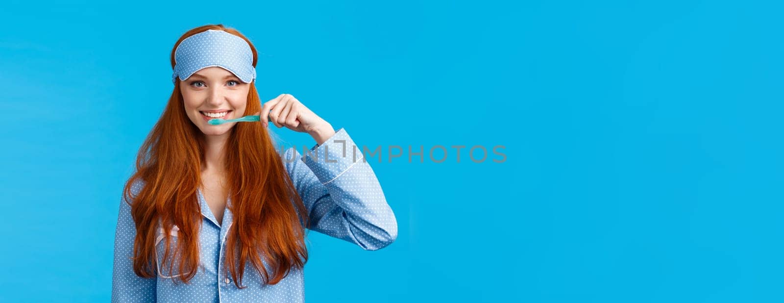 Habits, hygiene and lifestyle concept. Cute feminine redhead european woman in nightwear, sleep mask smiling and brushing teeth with toothbrush, standing blue background.