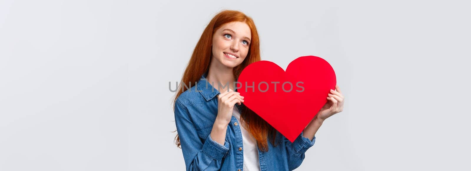 Dreamy, lovely caucasian redhead female dreaming to give valentines day card to lover, looking up thoughtful, imaging scene, holding big red heart and smiling happily, white background.