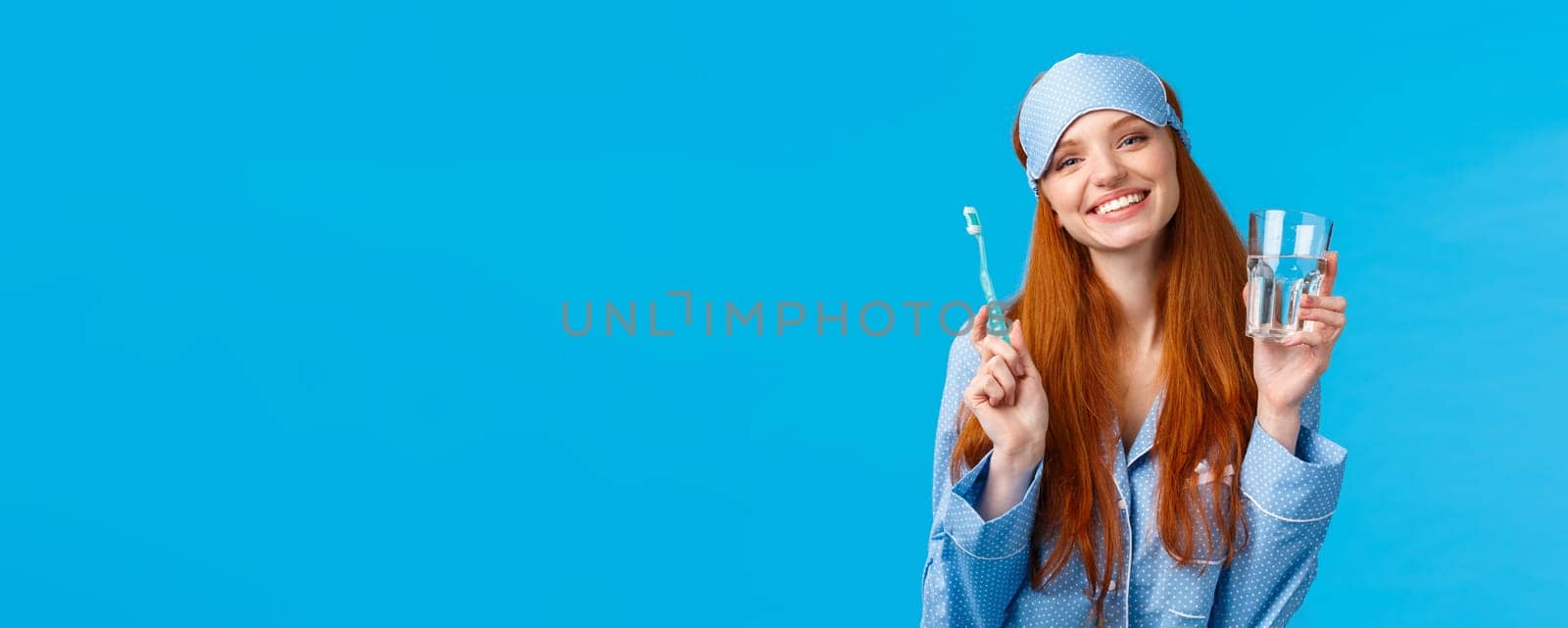 Happy smiling and carefree redhead enthusiastic girl in sleep mask, pyjama, holding glass water and toothbrush, taking care mouth hygiene, dental daily routine, blue background.