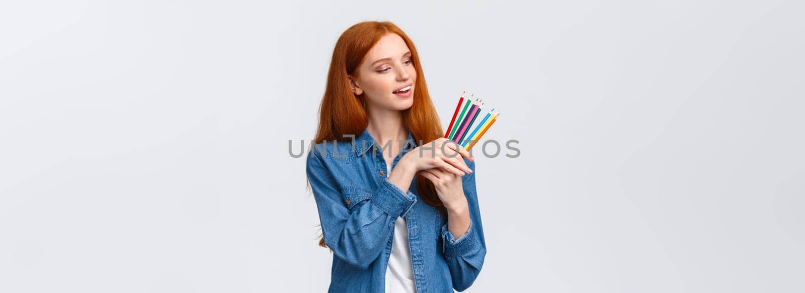 Creative and skilful good-looking redhead female in denim shirt, picking colored pencils, smiling thinking what draw, creating artworks, standing white background thoughtful.