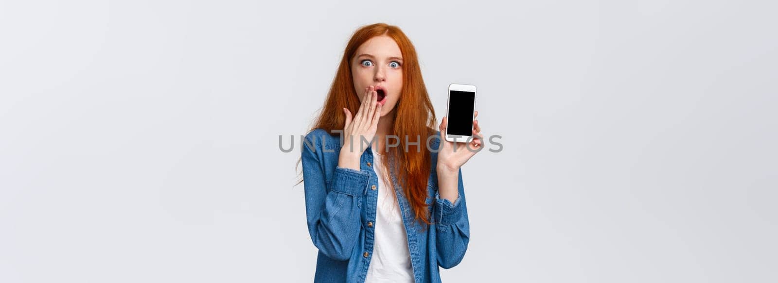 Impressed and astonished redhead woman seeing something unbelievable, spilling tea in internet, gasping cover opened mouth and showing samrtphone display, white background.