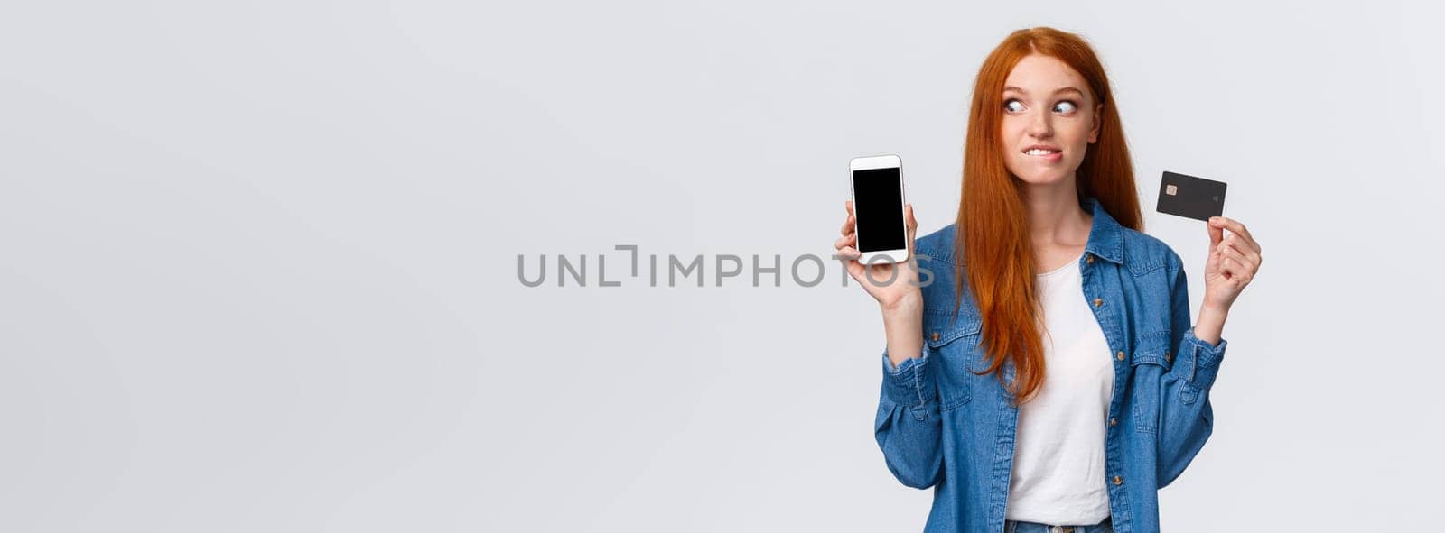 Shopaholic cant hold herself want buy something online. Eager and excited redhead girl see tempting price internet store, biting lip thrilled stare smartphone display, hold credit card.