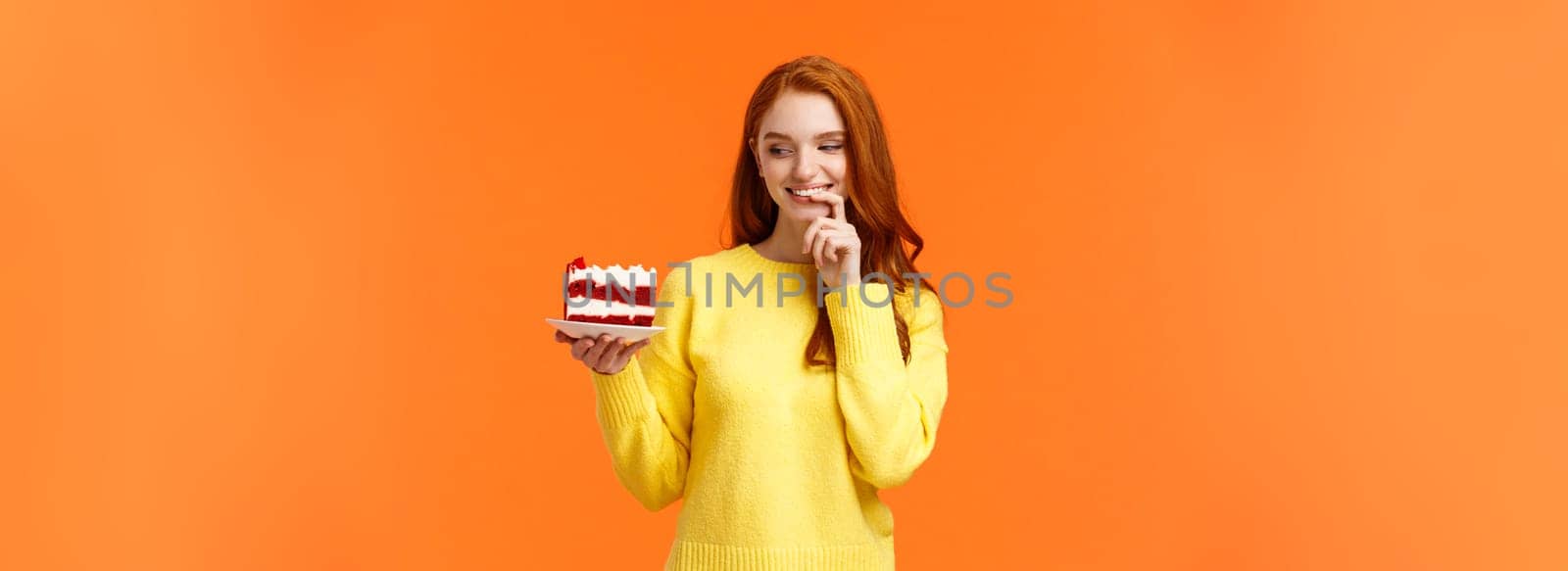 Cheerful and satisfied, happy redhead woman have cheat-day eating delicious food, holding tasty piece cake, biting lip and smiling, cant resist temptation, desire take bite, orange background.