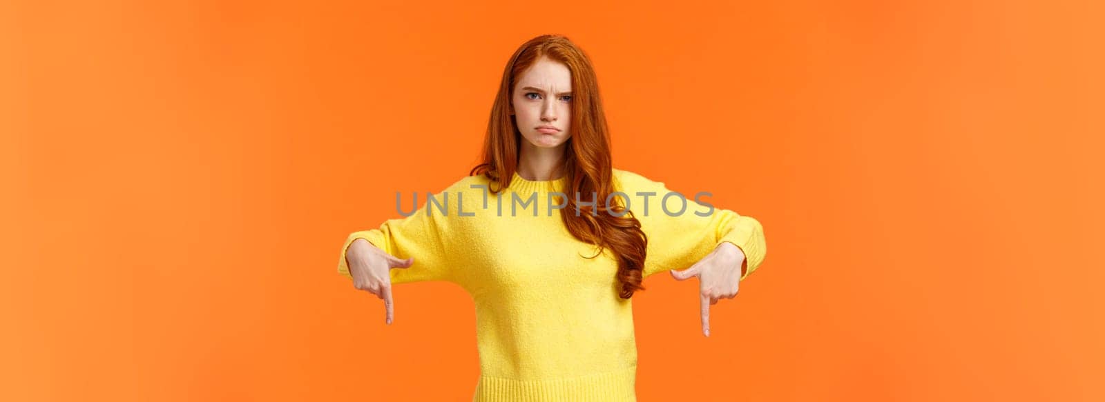 Mad unsatisfied cute redhead girlfriend, ginger girl in yellow sweater, frowning disappointed and angry, pointing down sulking, feeling offended with disapproval, standing orange background by Benzoix