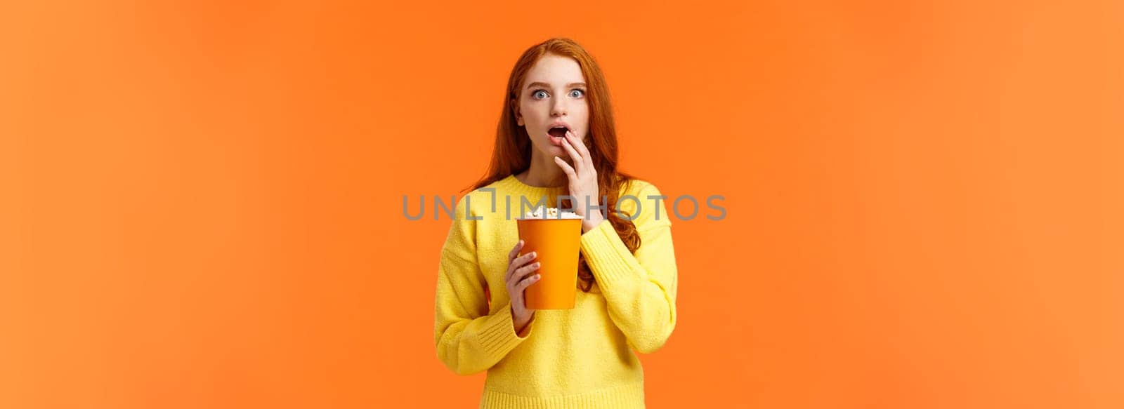 Shocked and interested, entertained redhead girl watching awesome new movie on big screen, eating popcorn, gasping open mouth astounded, startled of cool film plot, orange background.