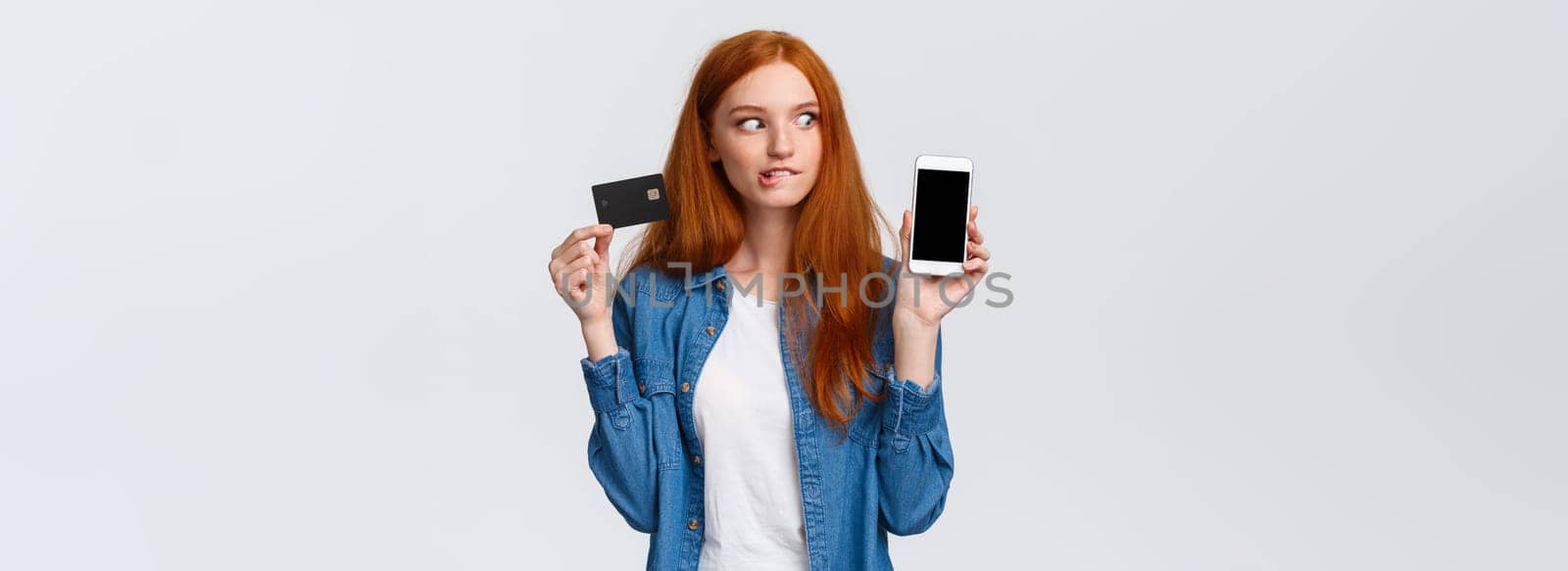 Girl consider buy thing with super good discount. Temped and wishful excited redhead female shopaholic looking at smartphone, shopping mobile app, holding credit card, bite lip eager purchase by Benzoix