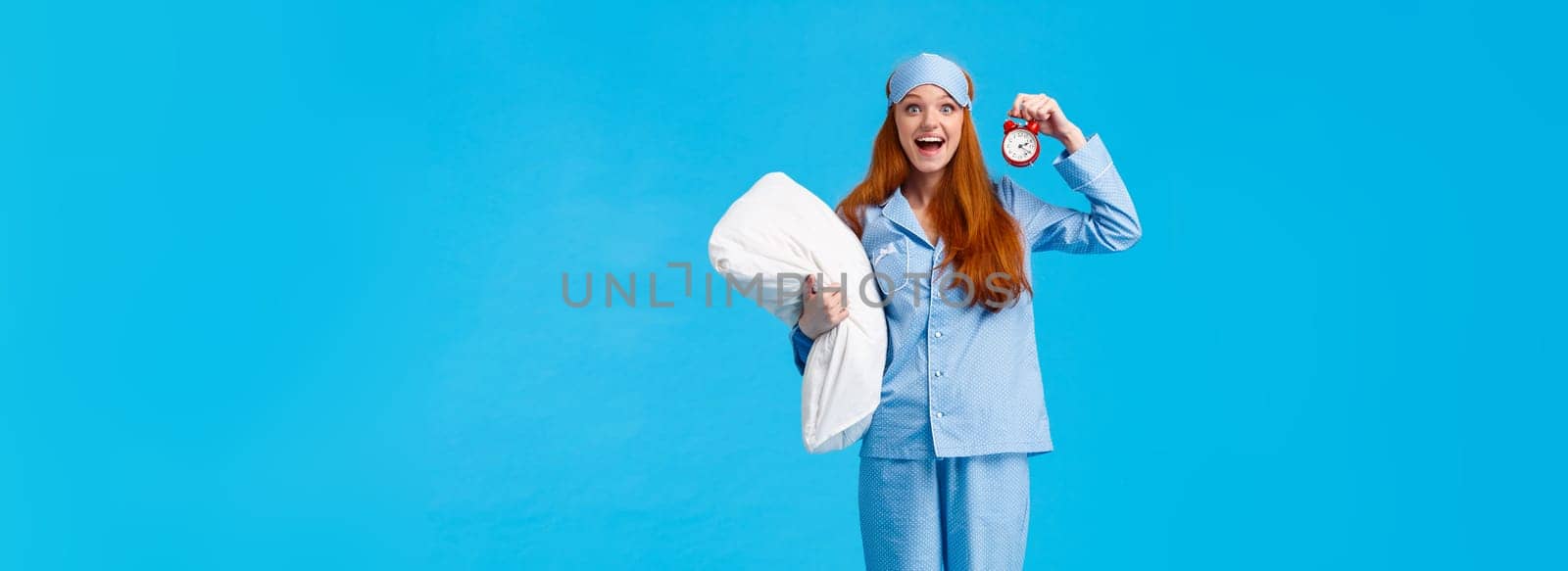 Full-length vertical studio attractive excited and happy cheerful upbeat girl in nightwear woke up energized in morning, holding red alarm clock, smiling taking pillow to bed, blue background.