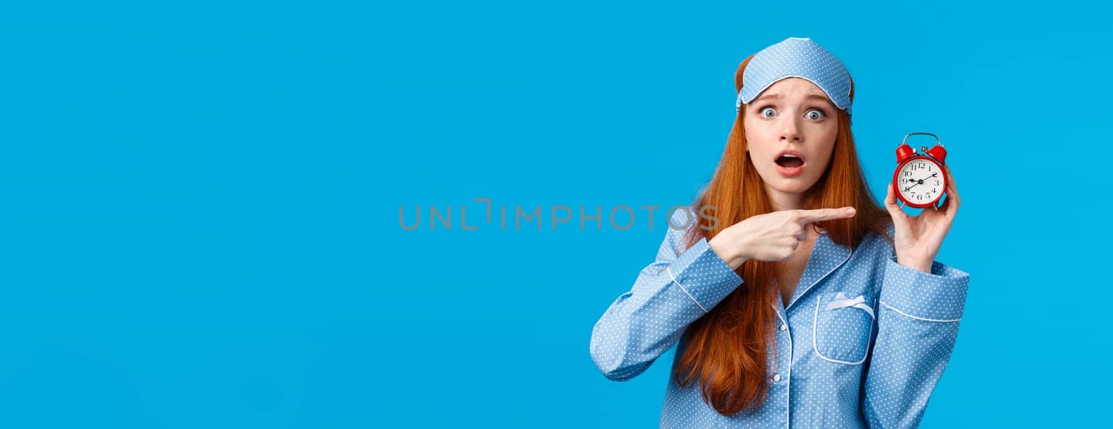Shocked and concerned, worried cute redhead girl pointing at alarm clock with frustrated, nervous expression, being late, dont know how set up right time, standing blue background in nightwear by Benzoix