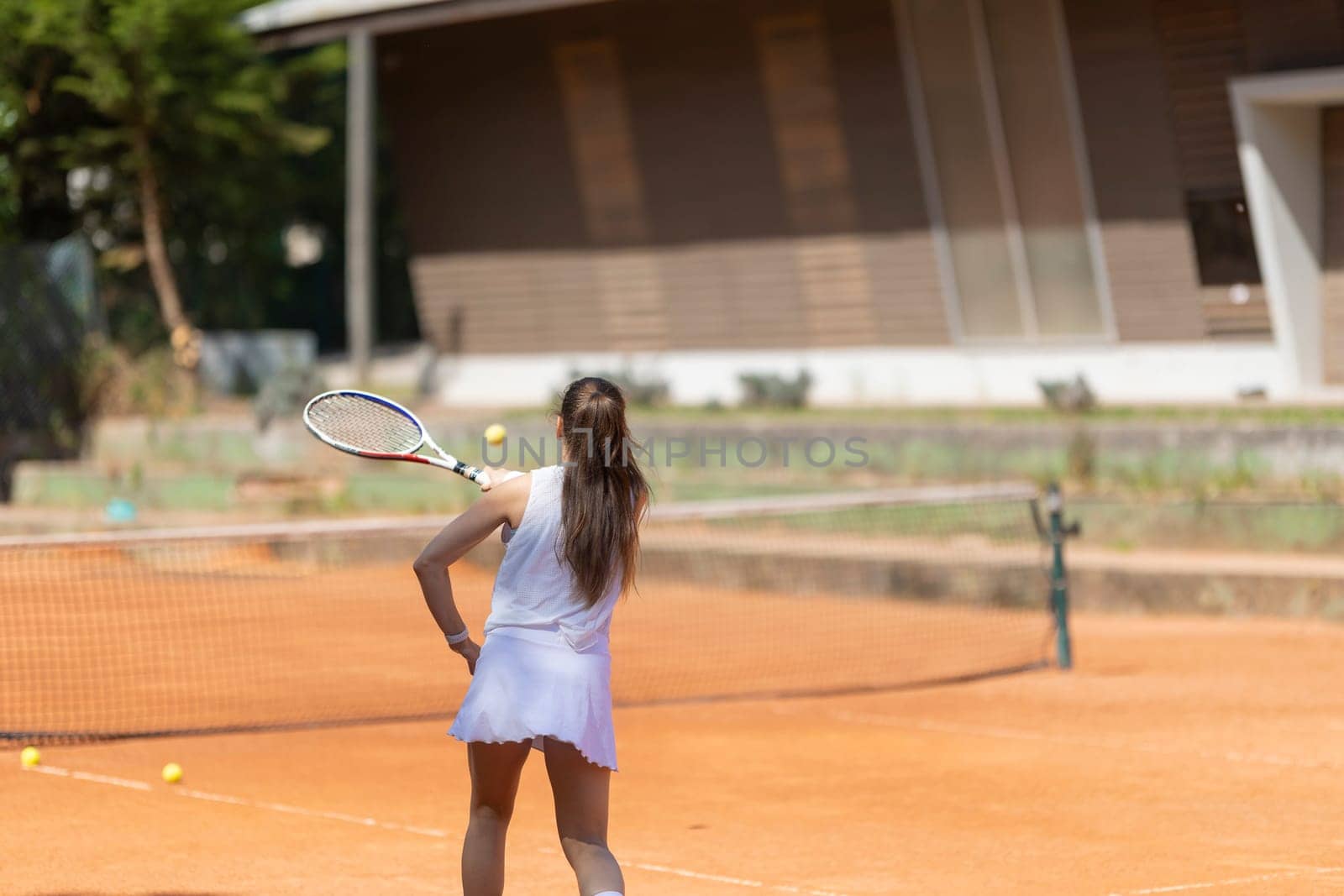 A woman is playing tennis on a clay court. She is wearing a white dress and holding a tennis racket