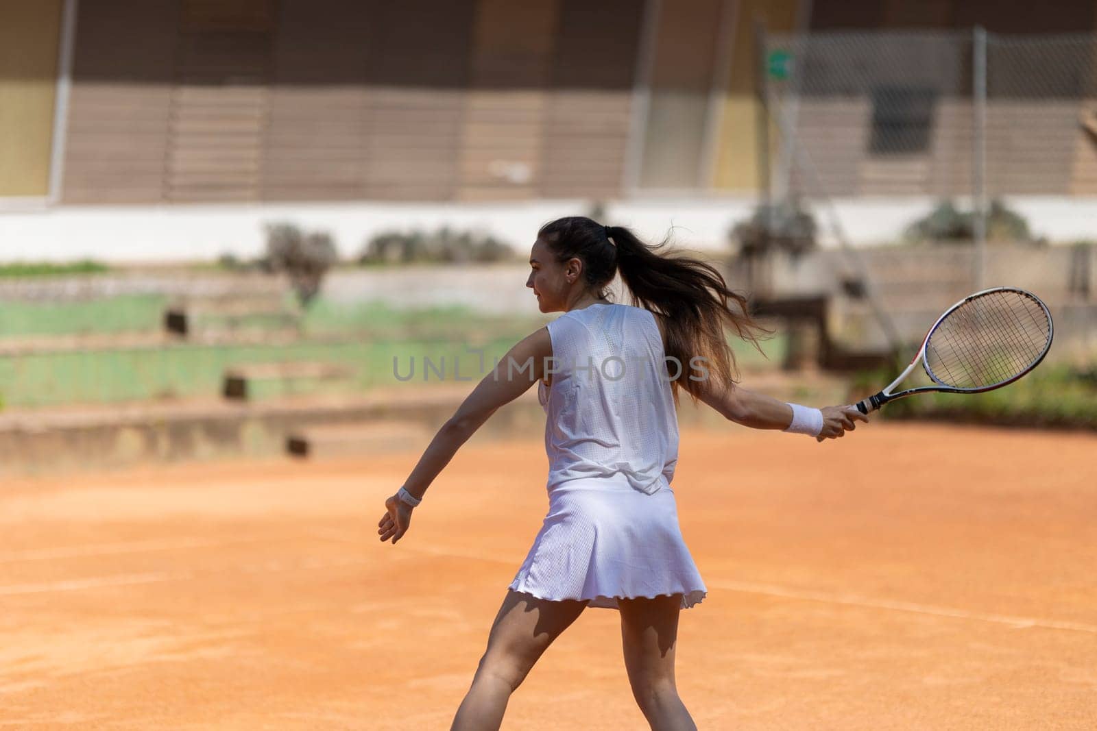 A woman is playing tennis on a clay court. She is wearing a white shirt and skirt and holding a tennis racket