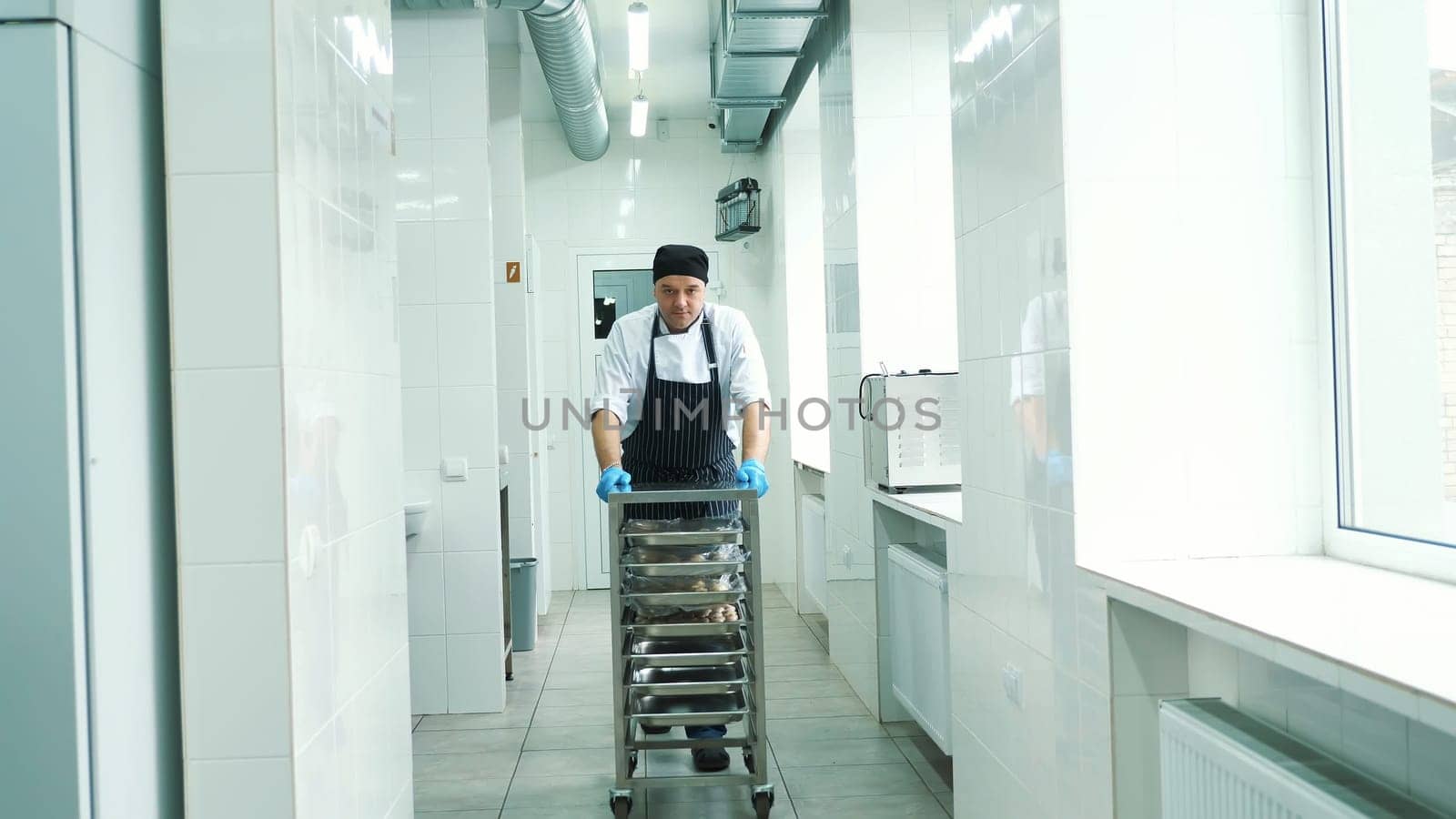 CHERKASY, UKRAINE, FEBRUARY 20, 2019: Commercial kitchen in canteen. A male cook walks through the kitchen hall, wearing an apron, carrying a special trolley for foodstuffs, blanks. High quality photo