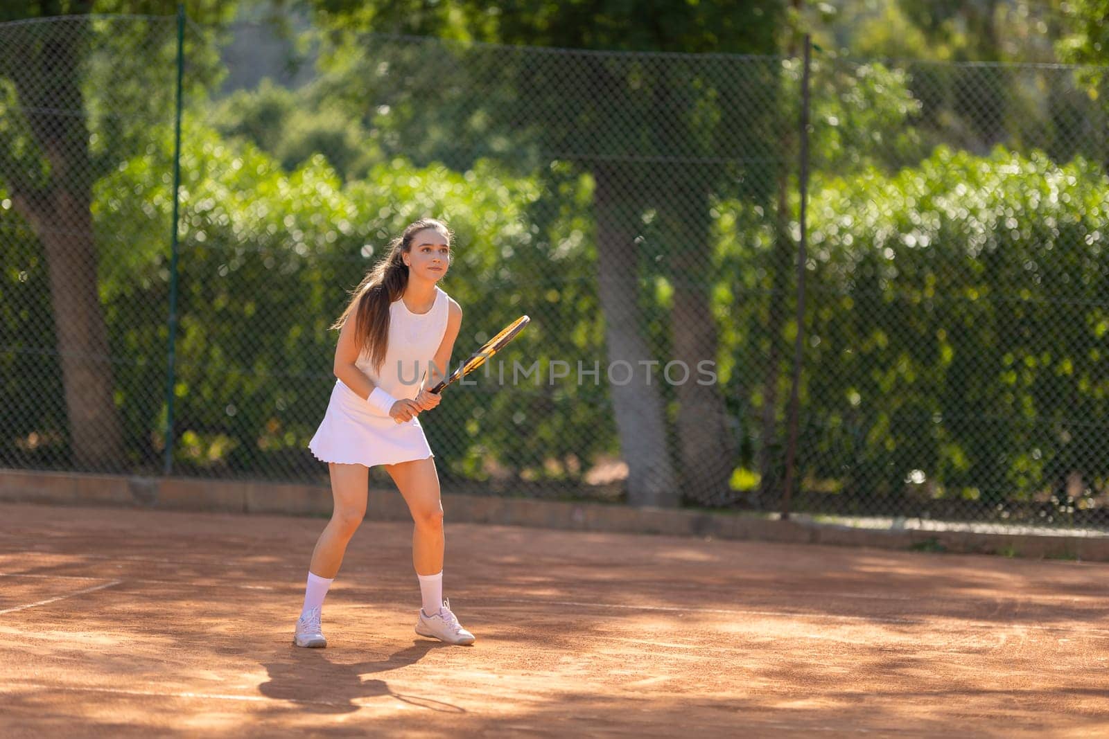 A woman is playing tennis on a clay court. She is wearing a white dress and is holding a tennis racket