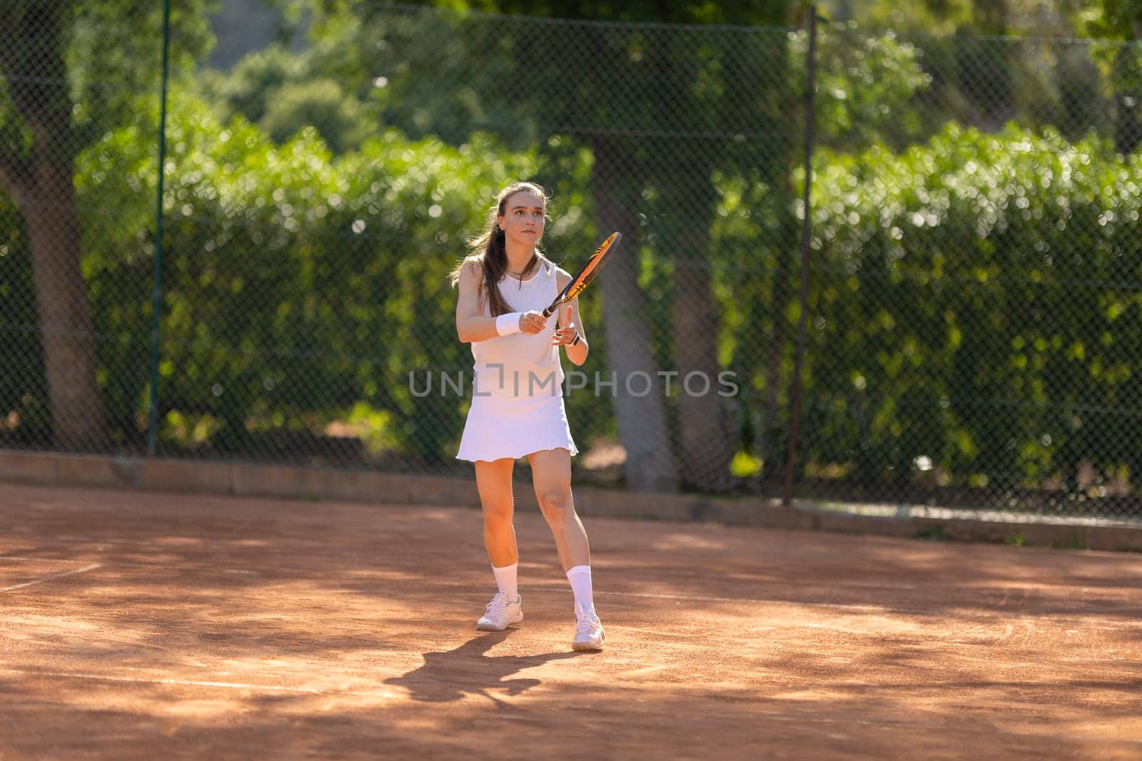 A young woman is playing tennis on a clay court. She is wearing a white dress and holding a tennis racket