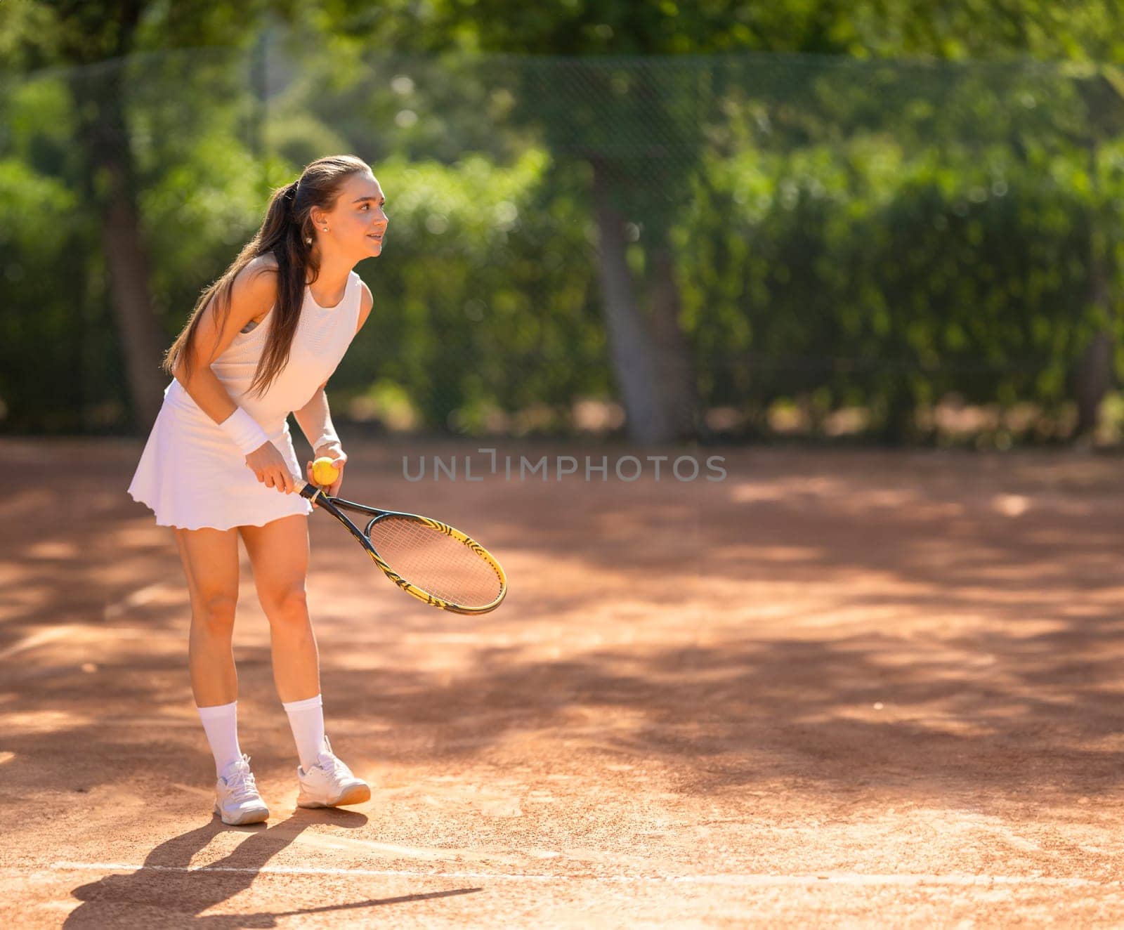 A woman is playing tennis on a clay court. She is wearing a white dress and a white tennis skirt
