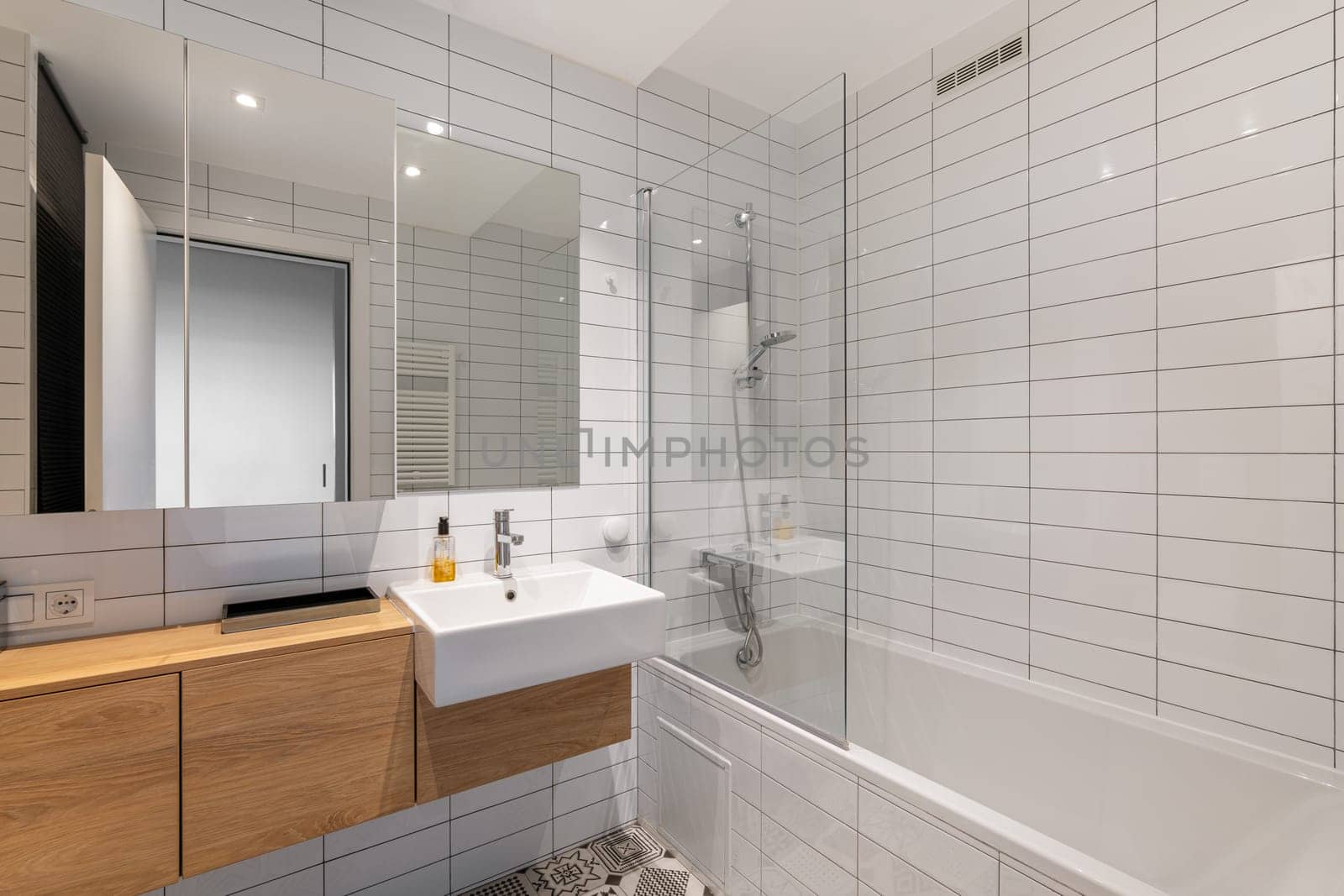 Clean and minimalistic modern bathroom featuring a wooden vanity, white tiles, a mirror, and a bathtub with glass enclosure.