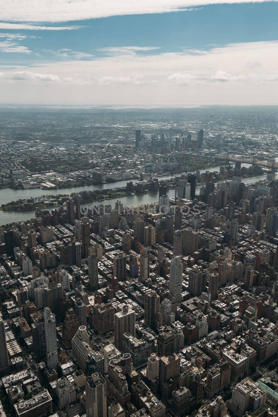 High-angle view of New York City showcasing numerous skyscrapers, the river, and vast cityscape under a partly cloudy sky. Image highlights the urban density and architectural diversity of the city.