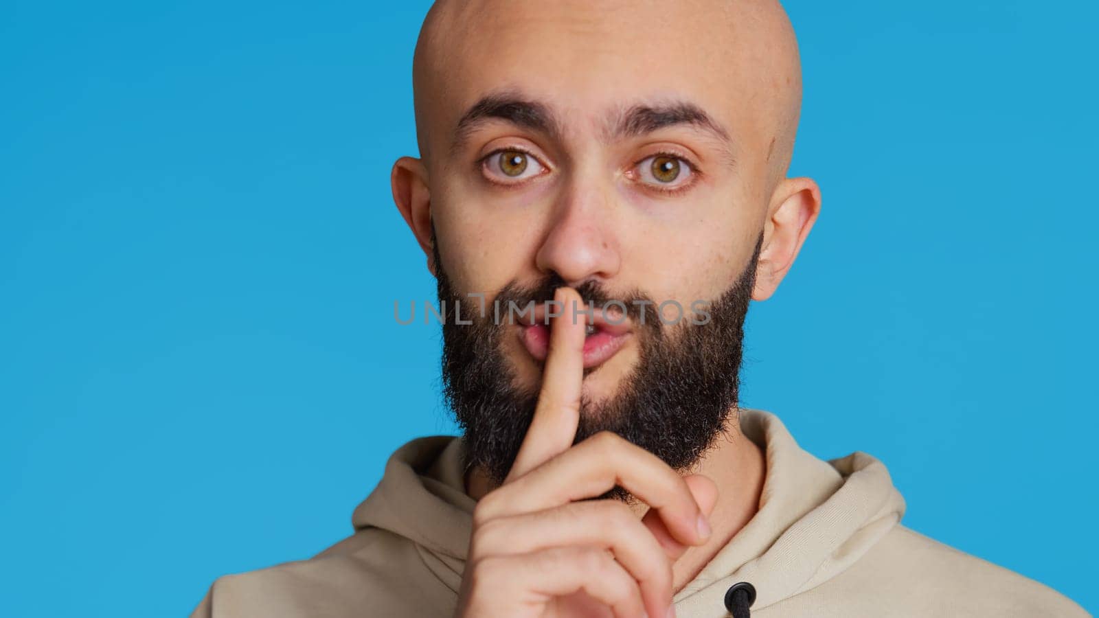 Middle eastern person showing mute gesture with finger over lips, posing over blue background. Arab guy doing hush symbol to keep silence, trying to share a secret whispering. Camera 2.