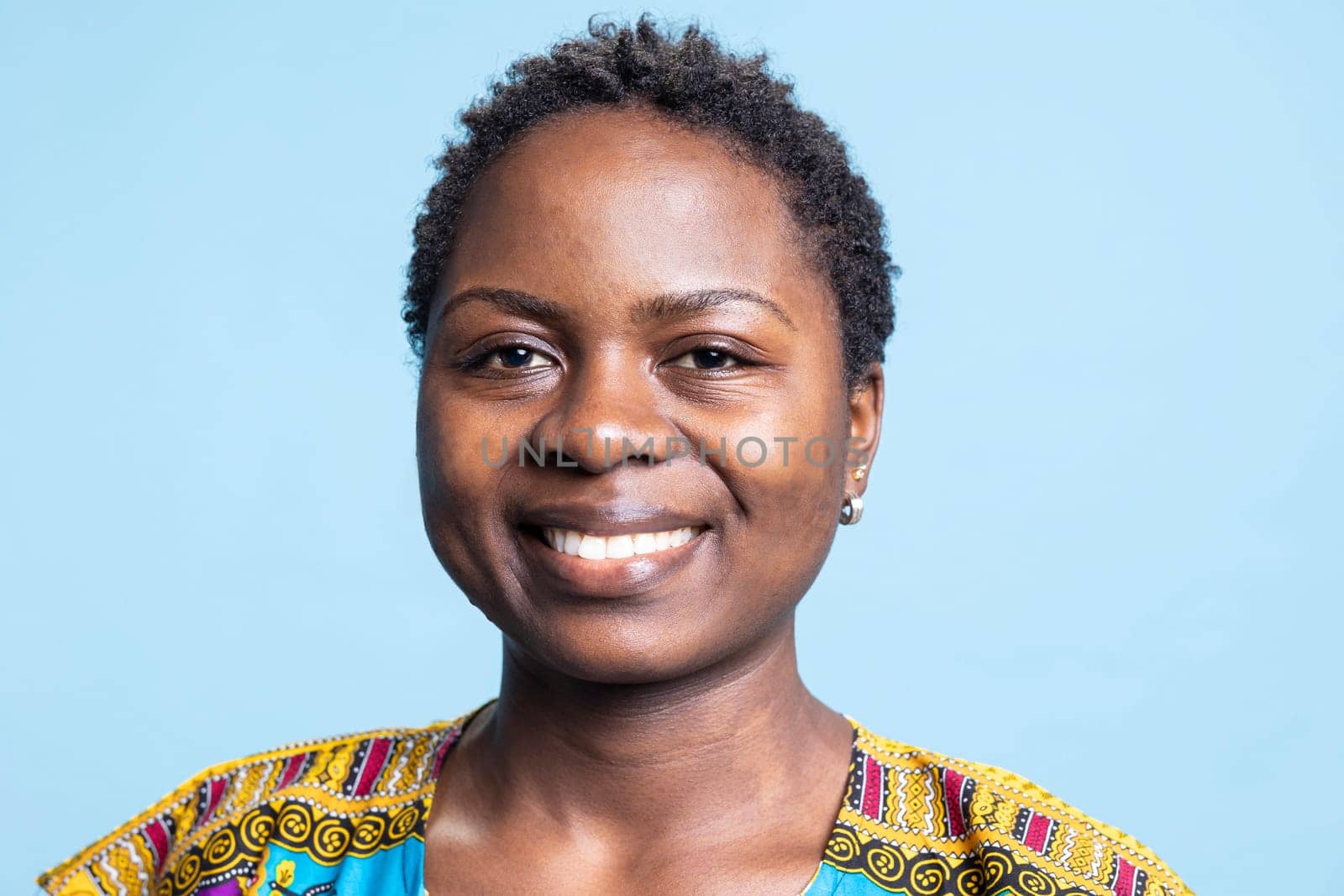 Portrait of cheerful female model posing over blue background, smiling while she wears traditional ethnic clothing. African american girl feeling confident in colorful outfit on camera.