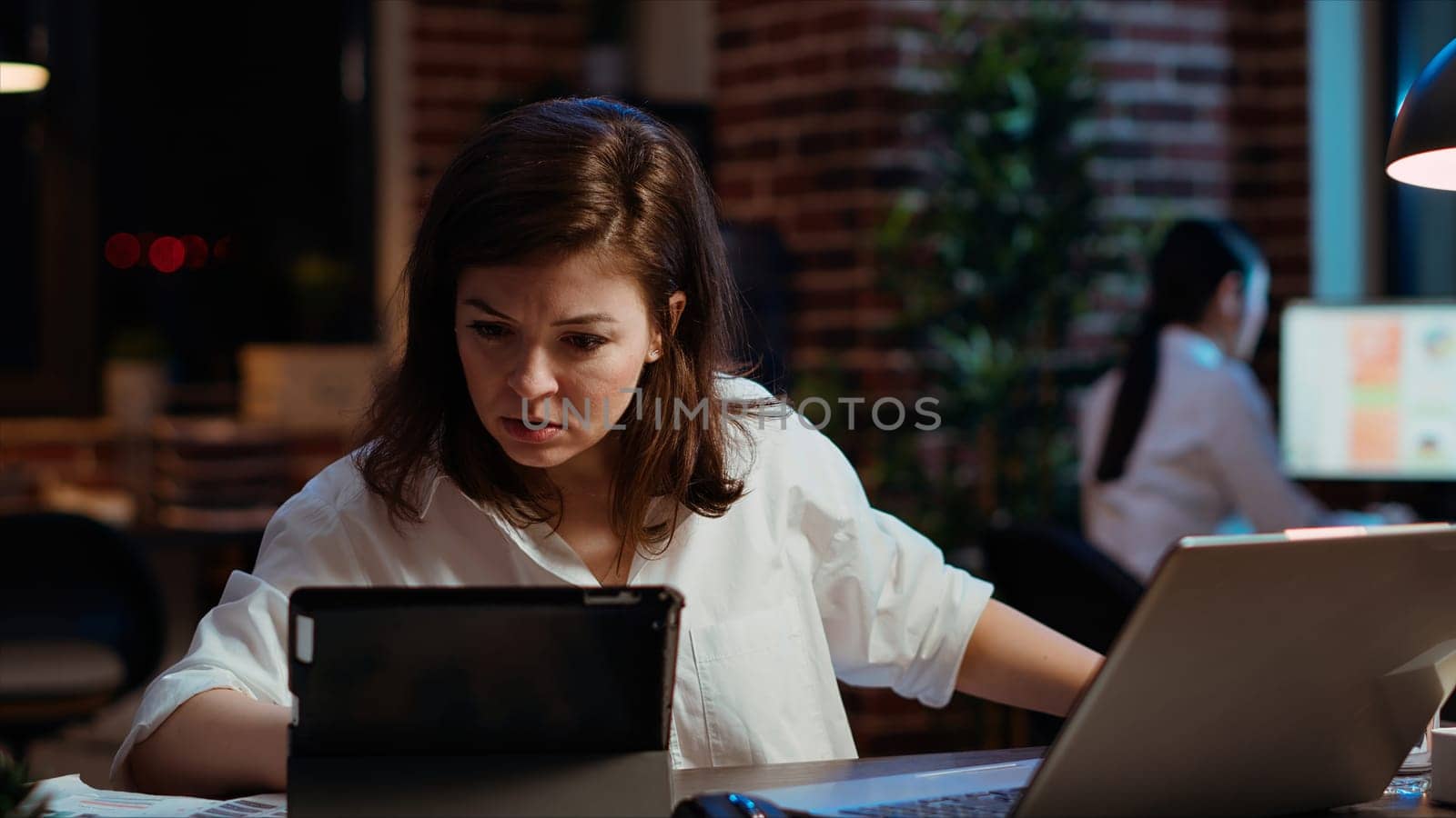 Worker looking over paperwork on tablet, crosschecking with data on laptop. Businesswoman looking over accounting figures on electronic devices late at night in brick wall office