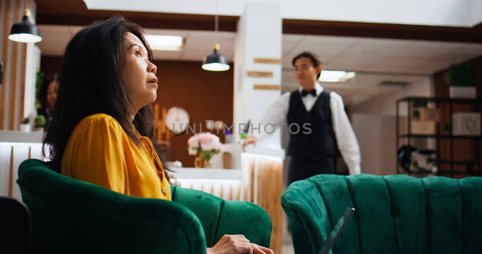 Hotel client solving issues on laptop in lounge area, waiting to register and see accommodation. Asian tourist passing time on pc, waiting to be called at front desk reception. Handheld shot.