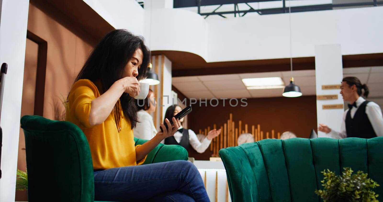 Tourist scrolling social media apps on smartphone, using free wifi connection in lounge area to pass time before doing check in. Asian woman relaxing on couch and planning trip itinerary.