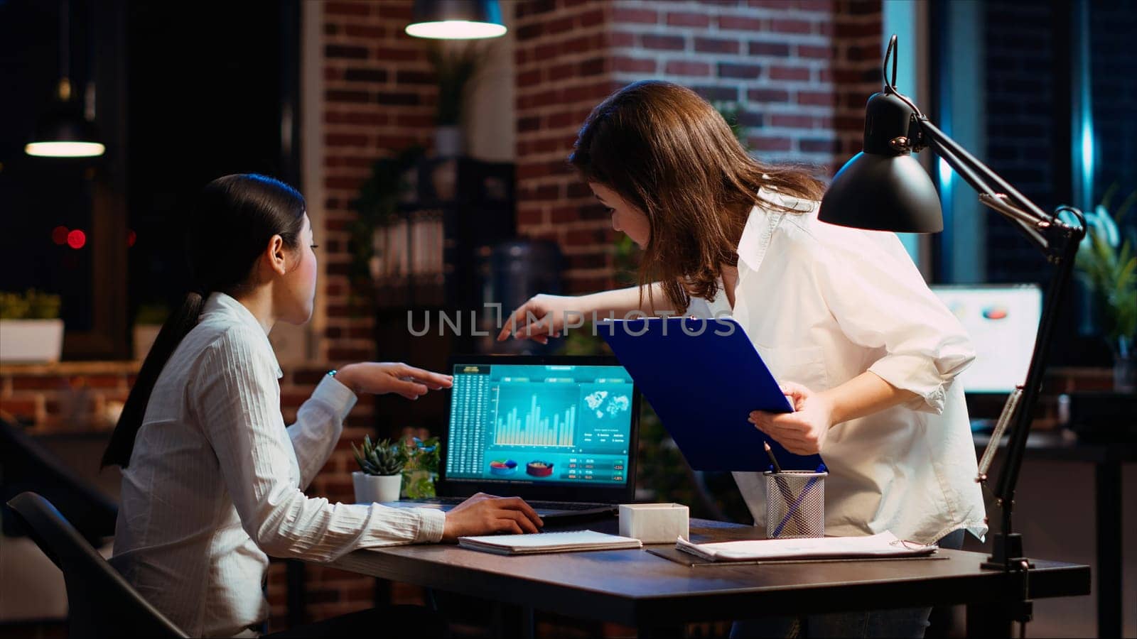 Company accountant and coworker analyzing financial data graphs on laptop screen. Employee and colleague comparing fourth quarter revenue figures in brick wall office, camera B