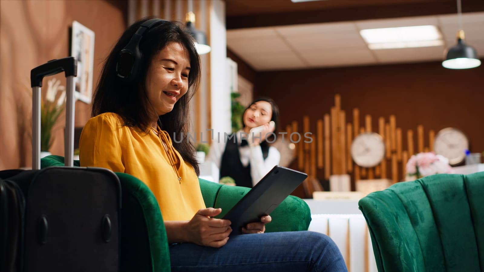 Asian tourist working on tablet in lounge area at resort, relaxing and waiting to confirm room reservation and do check in. Guest scrolling online websites on device, tourism concept. Handheld shot.