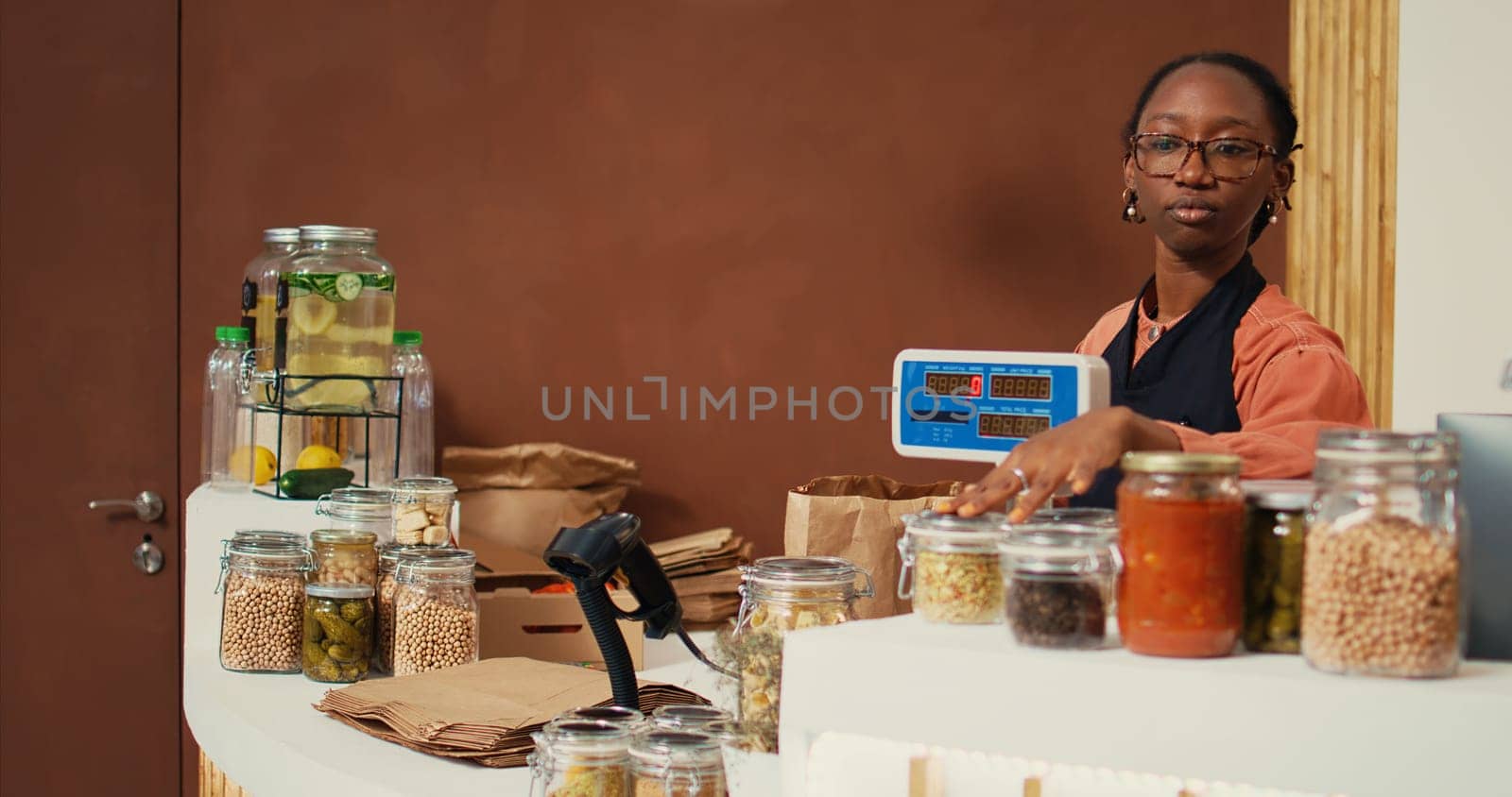 Retailer weighting goods on electronic scale at checkout, serving regular client with natural groceries from local supermarket. African american worker selling organic products. Handheld shot.