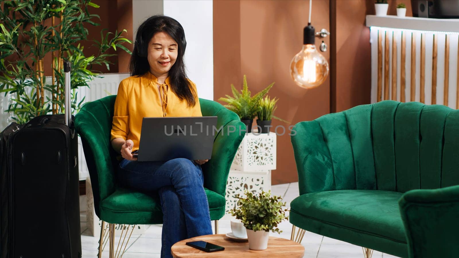 Asian tourist ordering coffee cup while she works on laptop, waiting for check in process in lounge area lobby. Woman arriving early at hotel, relaxing in foyer with refreshment.